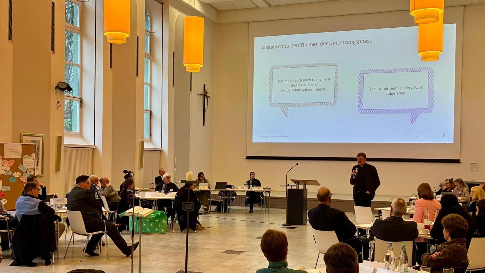 Ein Foto zeigt den Blick in einen großen Raum mit Leinwand. Personen sitzen Tischen und hören Kardinal Woelki zu.