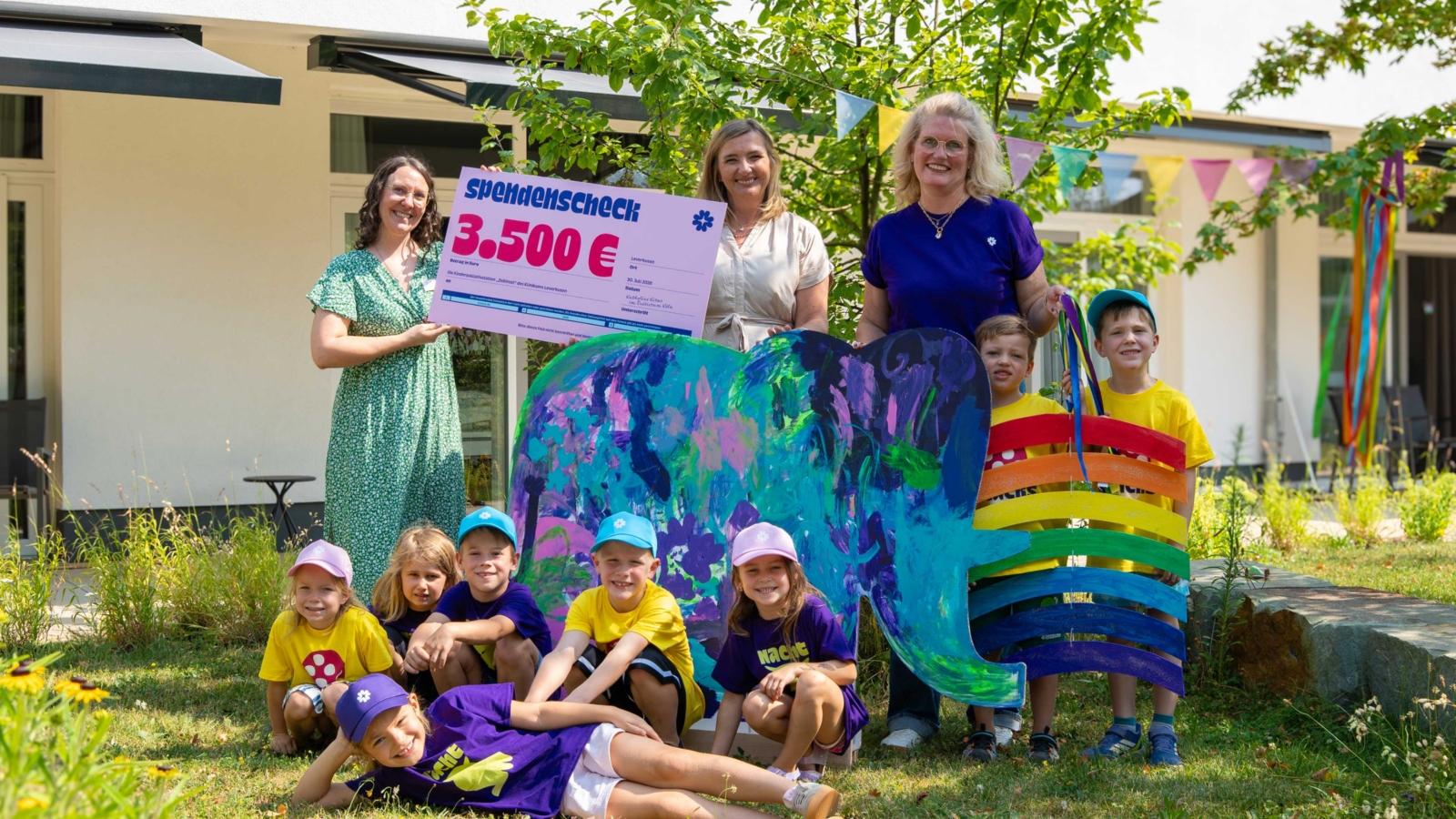 Agnes Busch, Geschäftsführerin von Katholino, überreicht gemeinsam mit Kindern der Kath. Kita St. Andreas den Spendenscheck und den beim Katholino-Sommerfest gestalteten Elefanten an Simone Vogel, Stationsleitung der Kinderpalliativstation „Zeitinsel“.